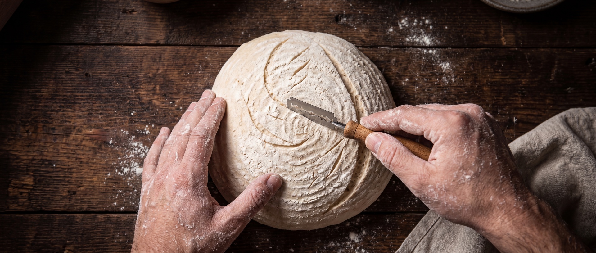 Baker scoring artisan bread dough on a wooden table.